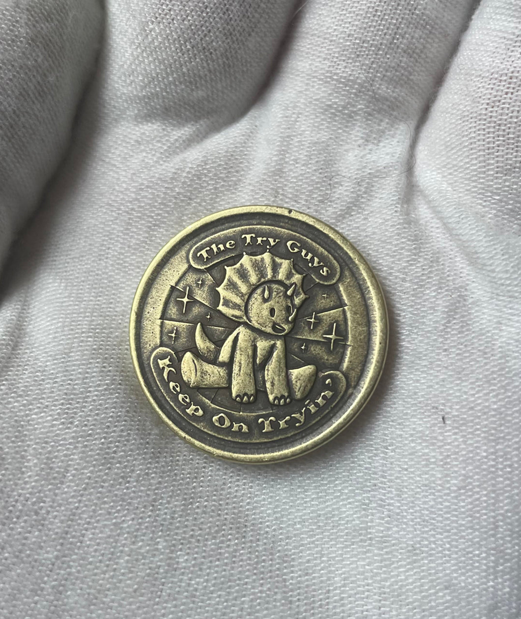 A gloved hand holds a brass coin. The coin has a struck design of a triceratops sitting and smiling. Around the triceratops is text that reads "The Try Guys" and "Keep On Tryin'". From the Try Guys.