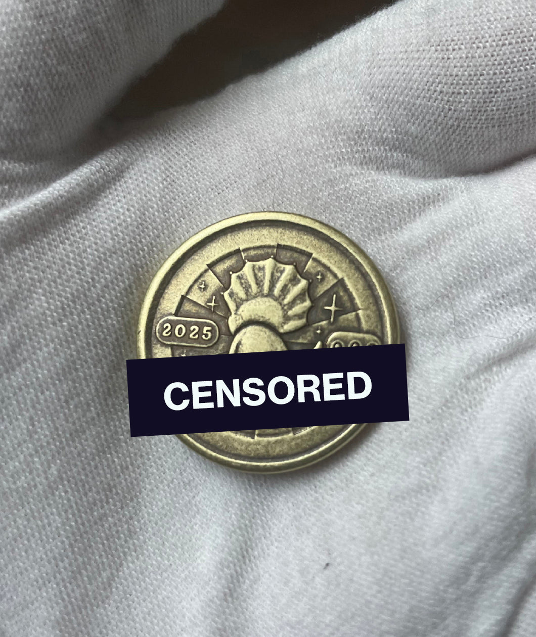 A gloved hand holds a brass coin. The coin has a struck design of a triceratops' back as it sits. Around the triceratops is text that reads "2005" and "000". Half of the coin is blocked out with a censor bar. From the Try Guys.