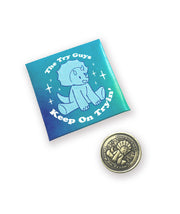 A folded envelope next to a brass coin. The envelope is meant to hold the coin and has a colorful design. The coin is brass and has a struck design of a triceratops and text that reads "The Try Guys" and "Keep On Tryin'". From the Try Guys.