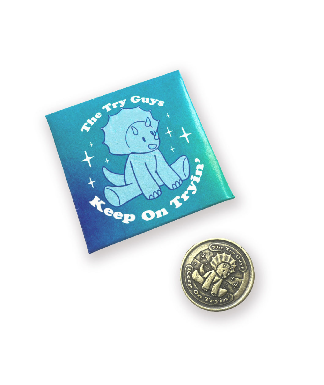 A folded envelope next to a brass coin. The envelope is meant to hold the coin and has a colorful design. The coin is brass and has a struck design of a triceratops and text that reads "The Try Guys" and "Keep On Tryin'". From the Try Guys.