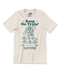 An off-white t-shirt with a design of a TV character waving printed in dark green on the front. Above the character is large text that reads, "Keep On Tryin'". Below the character are different versions of the character in tricky situations, like the tv guy slipping on a banana. Below those designs is text that reads, "Give The Try Guys method a try today!" From the Try Guys.