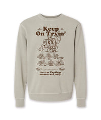 A tan crewneck sweatshirt with a design of a TV character waving printed in brown on the front. Above the character is large text that reads, "Keep On Tryin'". Below the character are different versions of the character in tricky situations, like the tv guy slipping on a banana. Below those designs is text that reads, "Give The Try Guys method a try today!" From the Try Guys.
