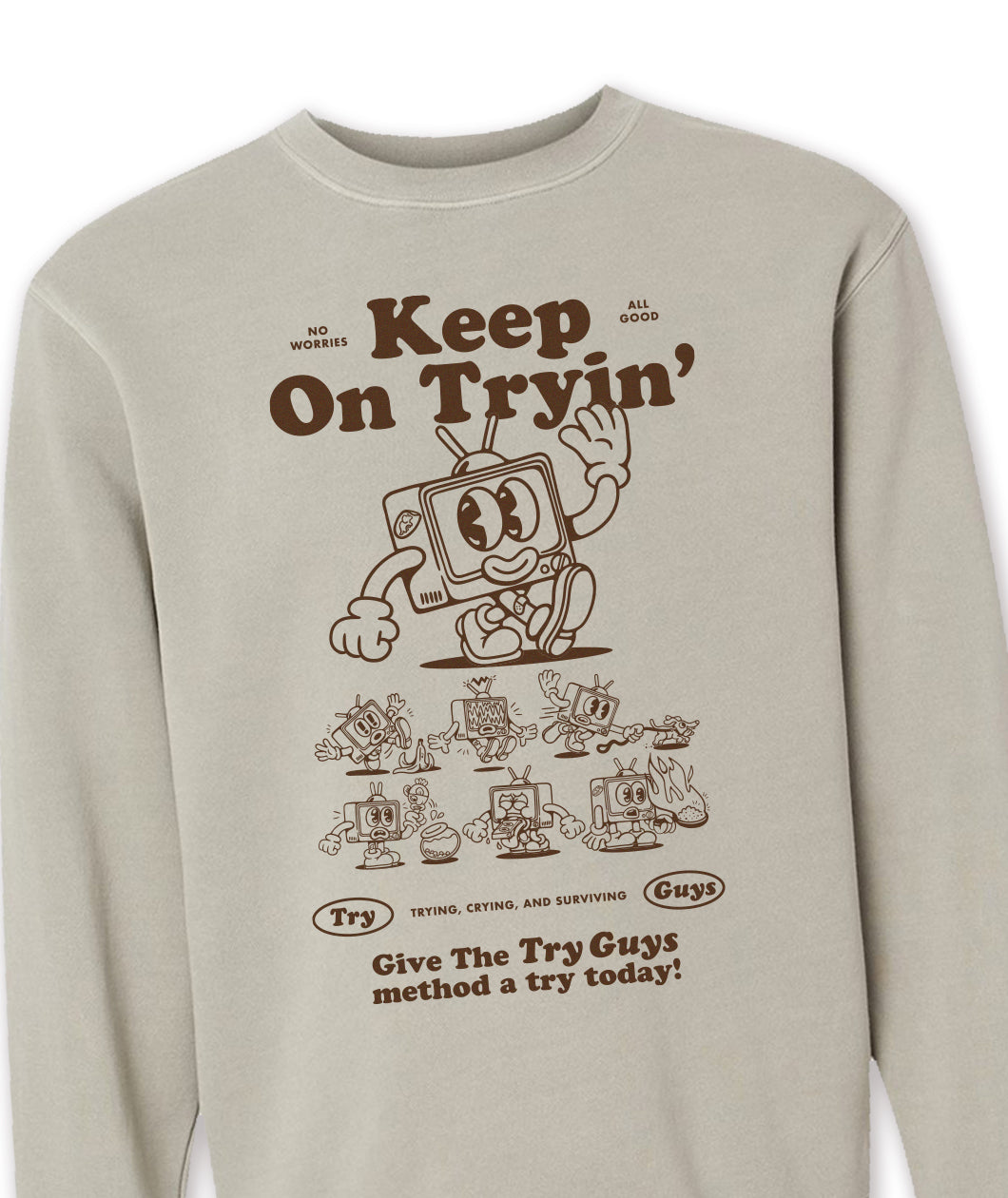 A zoomed-in tan crewneck sweatshirt with a design of a TV character waving printed in brown on the front. Above the character is large text that reads, "Keep On Tryin'". Below the character are different versions of the character in tricky situations, like the tv guy slipping on a banana. Below those designs is text that reads, "Give The Try Guys method a try today!" From the Try Guys.