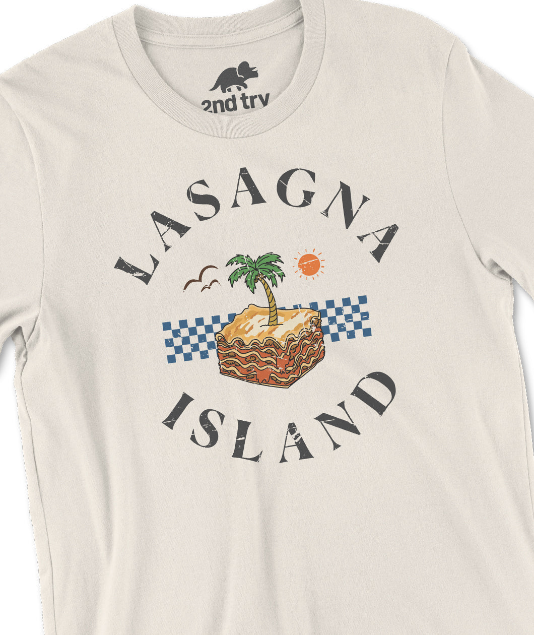 A closeup of an off-white t-shirt with a design of a piece of lasagna depicted as an island with a palm tree sprouting from its center. The lasagna is surrounded by birds and an orange sun in the distance. The design has text that reads, "Lasagna Island". From the Try Guys.