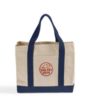 Front view of a canvas bag with navy bottom and handles featuring a front pocket with an embroidered patch with a red design of the Tryceratops logo and the text "The Try Guys"